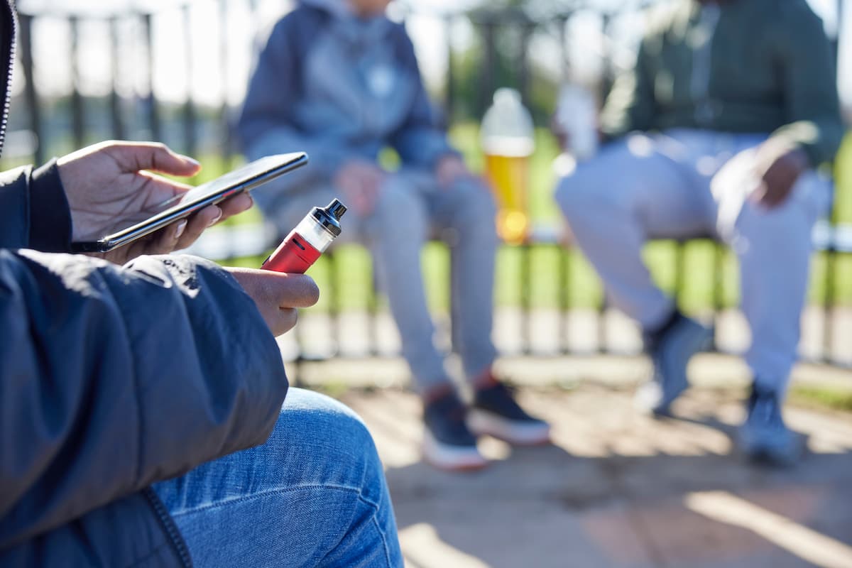 A photo of a few people sitting outside on benches in front of a school, with one of them on their phone and using a vape.